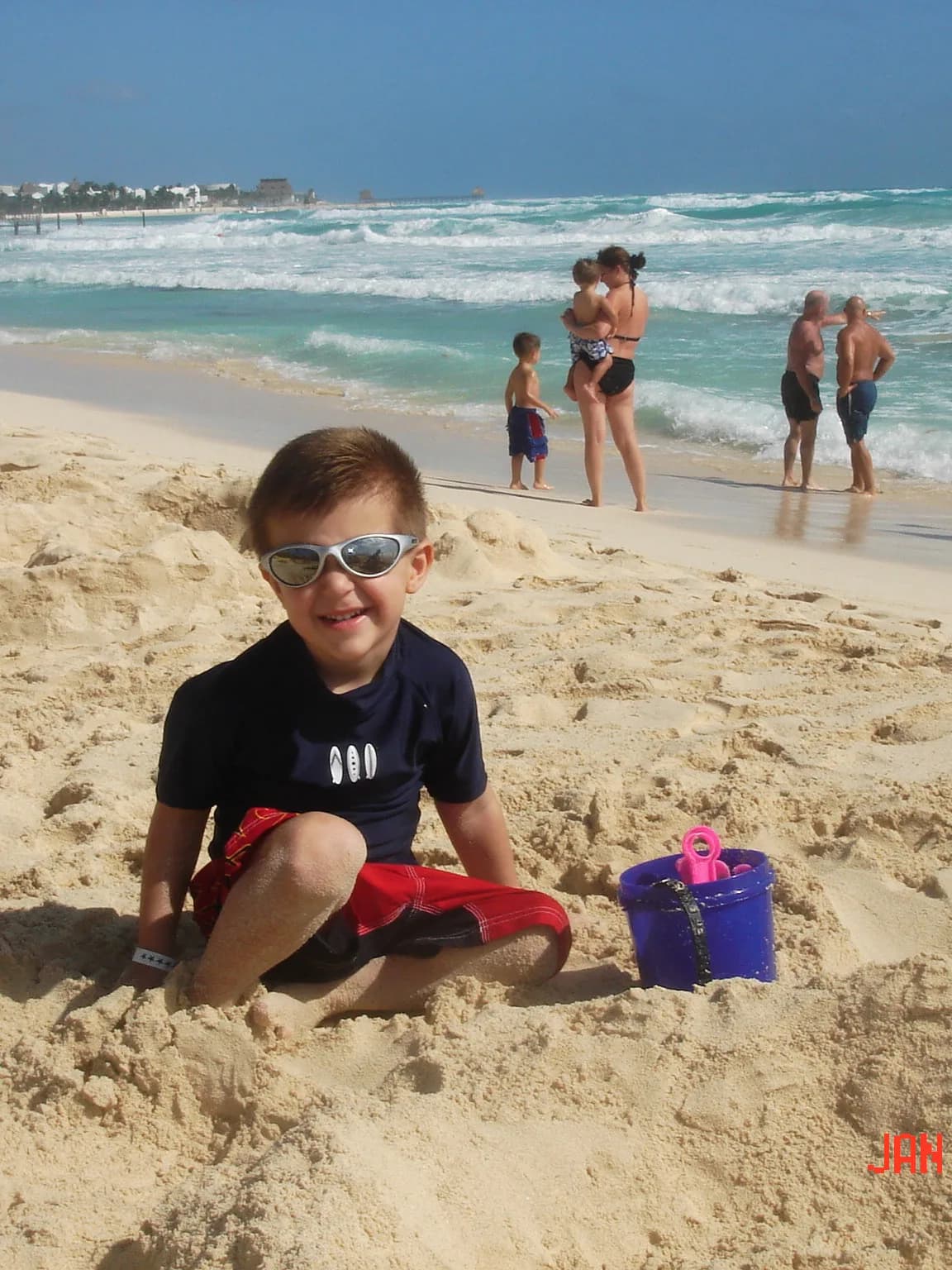 A child wearing sunglasses seated on the beach with a toy bucket